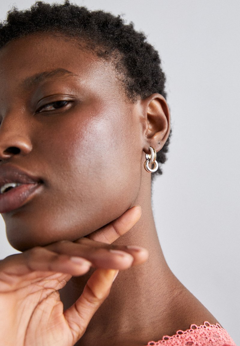 Cerceii din argint în formă de inel cu accente texturate; modelul prezintă un ton de piele natural, are părul scurt și ondulat, iar mâna este sprijinită pe maxilar, purtând un top coral.