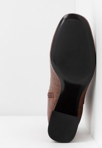 Chaussure en cuir marron avec texture crocodile, semelle plate noire et petit talon carré, reposant contre un mur d'angle blanc.