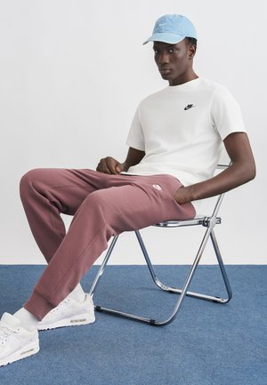 Noor mees istub metallist toolil, kandes valget Nike t-särki, tuhmi roosa Nike dressipükse, valgeid tossusid ja helesinist nokamütsi, taustaks ühevärviline sein.