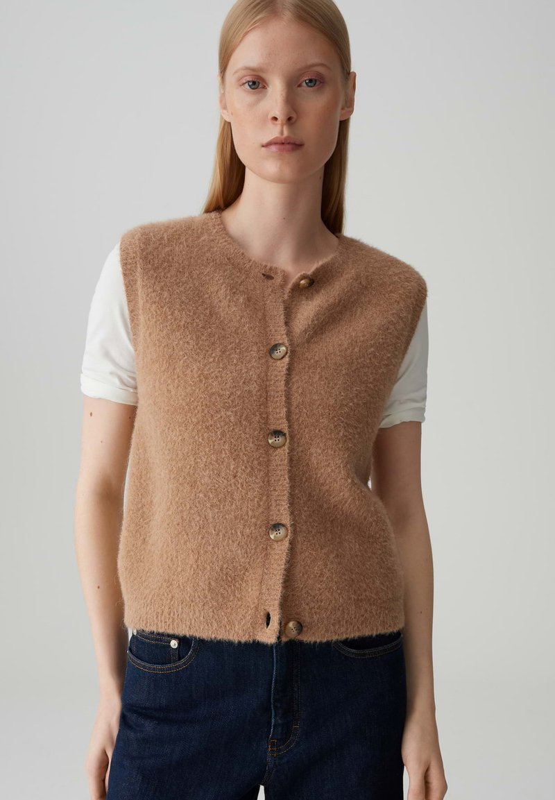 Gilet in misto lana beige con colletto rotondo, cinque bottoni frontali e una consistenza morbida e soffice; indossato sopra una camicia bianca.