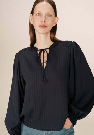 Blouse bleu marine avec un col en V et un détail à nouer. Présente des rayures verticales et des manches bouffantes avec des poignets froncés. Tissu léger.