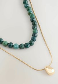 Zwei Halsketten auf einer hellen Oberfläche: eine mit facettierten grünen Perlen und eine dünne goldene Kette mit einem glatten, unregelmäßigen goldenen Anhänger.