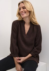 Blouse marron texturé avec un décolleté en V et des manches froncées, associée à un pantalon noir. Le design met en avant une silhouette décontractée et un matériau doux.