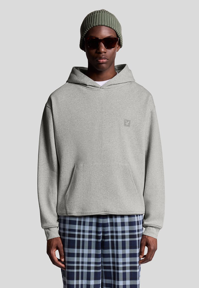 Uomo che indossa un berretto lavorato a maglia verde oliva, occhiali da sole tartaruga, felpa grigia chiara con tasca frontale e pantaloni a quadri blu su uno sfondo neutro.
