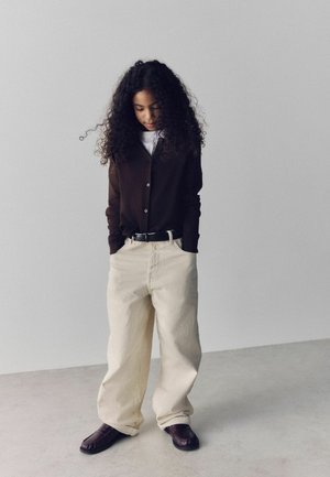Young person with curly hair wears dark brown cardigan, loose cream pants, black belt, and dark shoes, standing with hands in pockets.