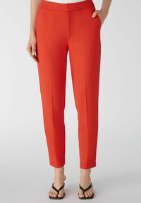 Pantalons rouges ajustés avec une texture lisse, dotés de poches latérales, d'un design à devant plat et d'une longueur de cheville effilée, associés à des sandales noires.