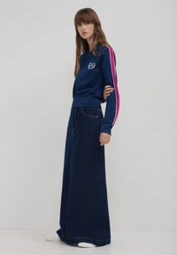 Giacca zip-up blu marina con strisce rosa e bianche sulle maniche, abbinata a una lunga gonna di jeans. Il modello indossa scarpe da ginnastica bianche e orecchini vistosi.
