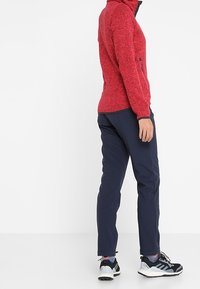 Personne portant une veste zippée rouge, un pantalon bleu marine et des chaussures de sport noires et blanches, debout devant un fond blanc uni.