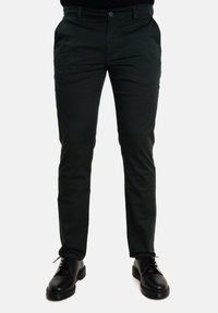 Pantaloni verde scuro con un design aderente, realizzati in tessuto liscio, dotati di tasche frontali e chiusura a bottone, abbinati a scarpe nere.