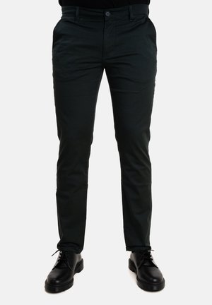 Pantaloni verde scuro con un design aderente, realizzati in tessuto liscio, dotati di tasche frontali e chiusura a bottone, abbinati a scarpe nere.