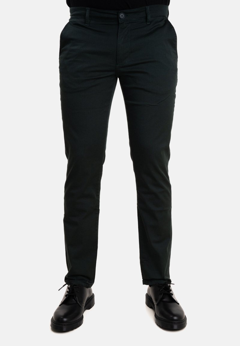 Pantaloni verde scuro con un design aderente, realizzati in tessuto liscio, dotati di tasche frontali e chiusura a bottone, abbinati a scarpe nere.