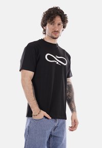 Uomo con capelli ricci che indossa una maglietta nera con un logo a forma di infinito con fulmine bianco, jeans blu e tatuaggi visibili sull'avanbraccio sinistro.