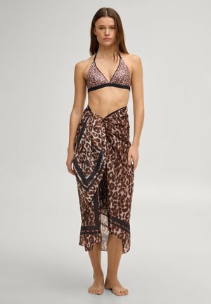 Woman wearing leopard print bikini top and matching sarong standing barefoot against plain background.