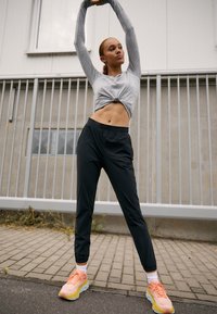 Tricou gri cu mâneci lungi și față legată, pantaloni negri tapered și sneakers roz cu accente galbene. Suprafață pavimentată și fundal gri.