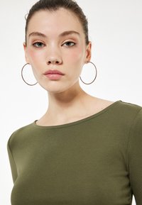 Olijfgroene getailleerde top met een ronde hals, geribbelde textuur en een gladde afwerking. Model draagt grote zilveren oorbellen.