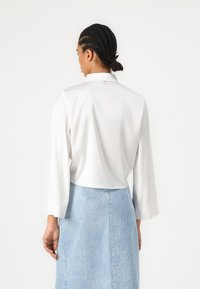 Blusa bianca con maniche lunghe e design corto, abbinata a una gonna di denim azzurro chiaro con cucitura centrale e una leggera sfumatura.