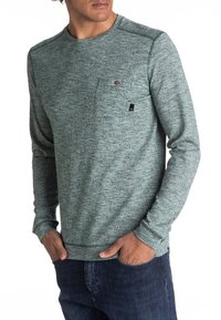 Felpa a maniche lunghe con collo a girocollo in tessuto teal testurizzato, dotata di taschino sul petto e piccolo logo del marchio. Abbinata a jeans scuri.