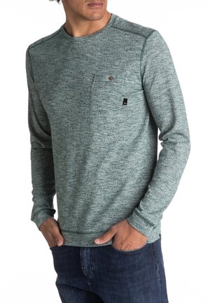 Felpa a maniche lunghe con collo a girocollo in tessuto teal testurizzato, dotata di taschino sul petto e piccolo logo del marchio. Abbinata a jeans scuri.