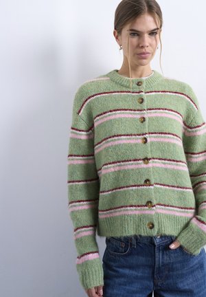 Ung kvinde iført grøn cardigan med knapper og pink og røde vandrette striber samt blå jeans, stående mod en ens hvid væg.