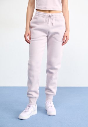 Vrouw draagt lichtroze joggerbroek, witte sneakers en een cropped top, staand op een blauwe vloer tegen een witte muur.