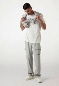 Hallid dressipüksid põlvetaskute ja tõmblukkudega, valge t-särgiga. Ta hoiab halli kampsunit üle õlgade. Valged tossud.