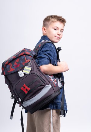 Boy in denim jacket carrying a large black and red backpack with reflective strips, standing against a plain white background.