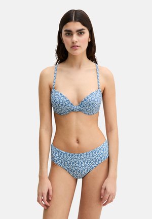 Młoda kobieta w niebieskim bikini z białym wzorem stokrotek, stojąca na jasnym jednolitym tle, zwrócona przodem.