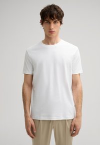 Camiseta de algodón blanca con cuello redondo, mangas cortas y un sutil logo bordado en el pecho, combinada con pantalones beige.