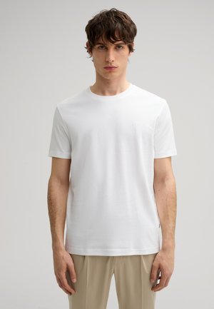 Weißes Baumwoll-T-Shirt mit Rundhalsausschnitt, kurzen Ärmeln und einem dezenten gestickten Logo auf der Brust, kombiniert mit beigefarbenen Hosen.