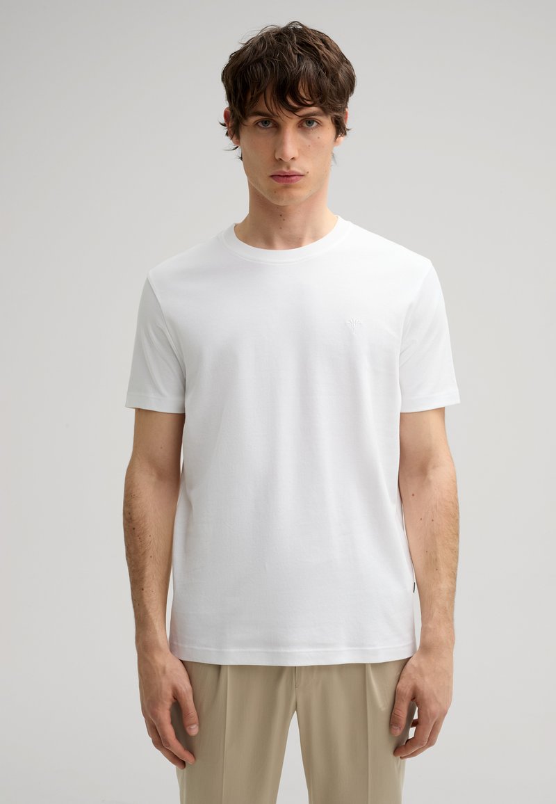 Camiseta de algodón blanca con cuello redondo, mangas cortas y un sutil logo bordado en el pecho, combinada con pantalones beige.