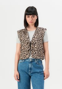 Jonge vrouw met stijl zwart haar draagt een mouwloze vest met luipaardprint over een grijze T-shirt en hooggewaadde blauwe jeans, staande tegen een witte achtergrond.