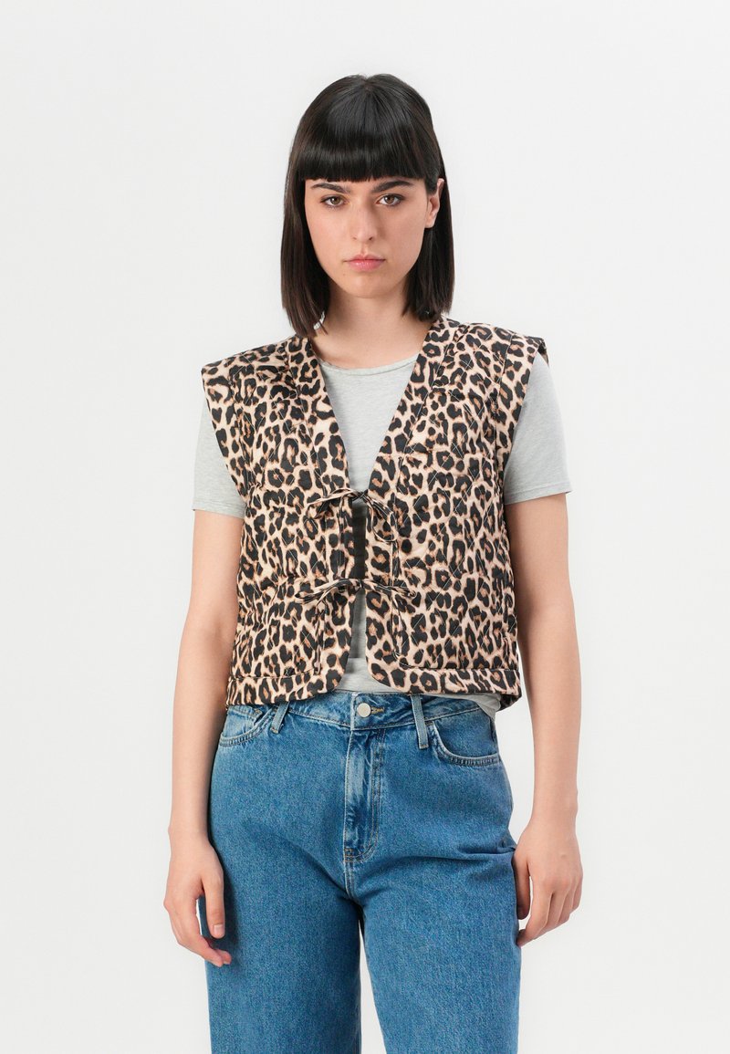 Jonge vrouw met stijl zwart haar draagt een mouwloze vest met luipaardprint over een grijze T-shirt en hooggewaadde blauwe jeans, staande tegen een witte achtergrond.