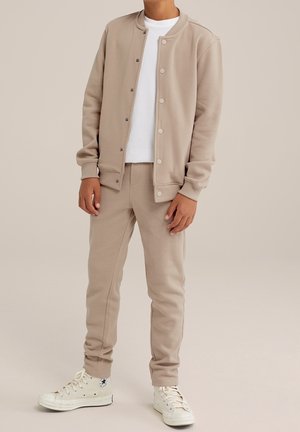 Beżowy sweatshirt z zapięciem na napy, w zestawie z pasującymi beżowymi spodniami dresowymi i białą koszulką; strój charakteryzuje się miękką fakturą i luźnym krojem.