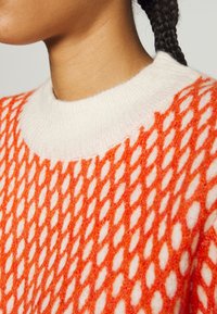 Pull en tricot orange avec un motif en diamants texturé, présentant un col rond blanc côtelé. Gros plan sur les mailles et le contraste des couleurs.