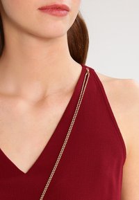 Bordowa bezrękawnik z głębokim dekoltem w kształcie litery V, teksturowany materiał i złoty łańcuszek na ramieniu, podkreślający detale projektu.