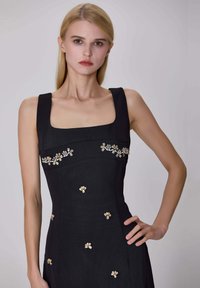 Femme jeune avec des cheveux blonds raides, portant une robe noire sans manches avec un décolleté carré et de petites embellissements floraux, posant avec une main sur la hanche.