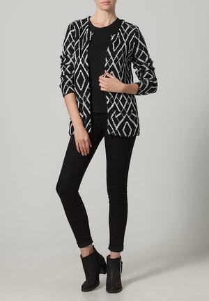 Cardigan a motivi in bianco e nero con apertura frontale, maniche lunghe e design a rombi, abbinato a un top nero e jeans skinny, insieme a stivali alla caviglia.