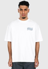 White cotton t-shirt with short sleeves, featuring a blue graphic print on the left chest. Relaxed fit and crew neckline.