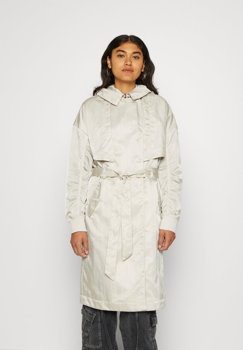Trench coat de satén beige claro con capucha, cintura ceñida y cierre frontal de botones. Cuenta con mangas largas y bolsillos laterales.