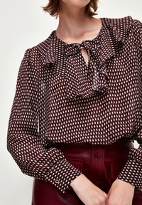 La blusa presenta una base color burdeos con un estampado blanco y azul, cuello y puños con volantes, y una abertura en forma de llave en el escote. Corte estructurado.