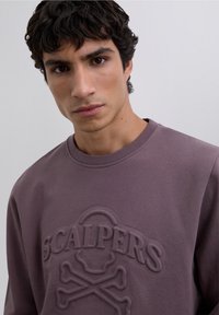 Sudadera púrpura con logo "SCALPERS" en relieve y gráfico de calavera y huesos cruzados. Fabricada en tela suave con cuello redondo.