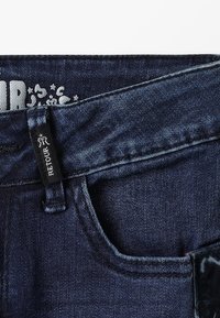 Jeans de mezclilla azul oscuro con un diseño clásico y una etiqueta visible en la cinturilla. Textura suave y colocación estándar de los bolsillos.