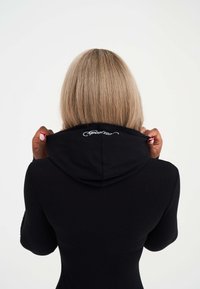 Schwarze, kapuzenähnliche Kleidung mit gerippter Textur. Die Rückansicht zeigt Hände, die die Kapuze halten. Verfügt über ein dekoratives weißes Logo im Nackenbereich.