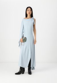 Ανοιχτό μπλε φόρεμα με έναν ώμο και λεπτομέρεια κορδέλας, διαθέτοντας πλάγιο σκίσιμο. Συνδυασμένο με μεταλλική τσάντα χειρός και μαύρες μπότες με τακούνι.
