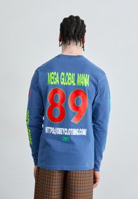 Camisa de manga larga azul con gráficos grandes: "MEGA GLOBAL MAMA" y "89" en rojo y verde. Mangas con detalles textuales.
