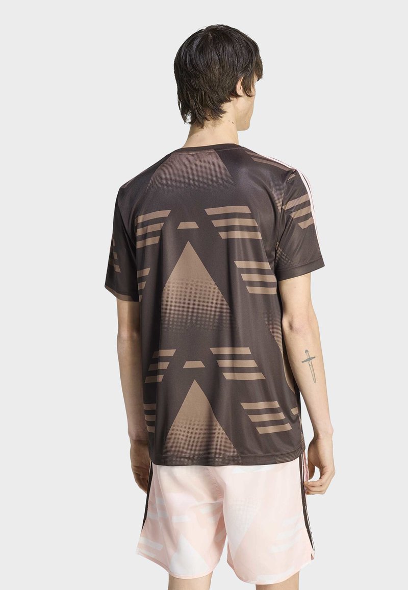 Persona con capelli corti e scuri vista di spalle, che indossa una camicia marrone con motivo geometrico e pantaloncini rosa chiaro, con un tatuaggio di spada sul braccio sinistro.