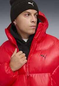 Rode pufferjack met een glanzende afwerking, voorzien van een zwart Puma-logo. Gedeeltelijk over een zwarte hoodie gedragen, de stof is gewatteerd en gestikt.