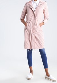 Ljusrosa lättviktig jacka med huva, framdragkedja och sidofickor, parat med blå ankel-långa jeans och vita slip-on skor.