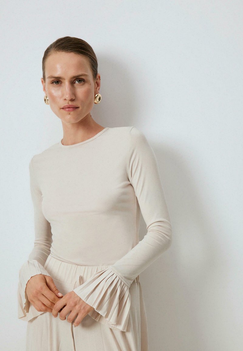 Žena oblečená v béžovom dlhom rukáve s plisovanými zvonovými manžetami a zladenými vysokými nohavicami, doplnená veľkými okrúhlymi zlatými náušnicami.