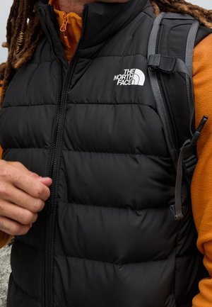 Osoba nosząca czarną pikowaną kamizelkę North Face na pomarańczowej bluzie z kapturem, z widocznymi paskami plecaka na ramieniu.